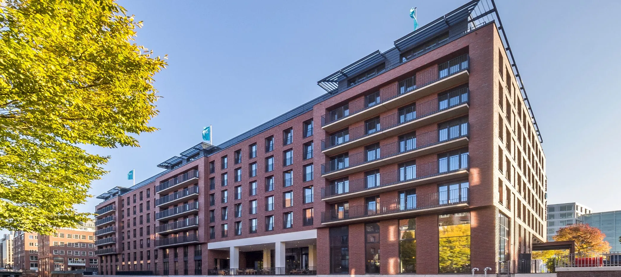 Das Bild zeigt das Motel One in der Admiralstraße in Hamburg, das direkt an einem kleinen Fluss liegt. Das Gebäude ist in rote Klinker gehüllt, was ihm eine markante, traditionelle Ausstrahlung verleiht. Die Sonne strahlt am blauen Himmel und wirft ihre warmen Strahlen auf das Motel. Besonders hervorzuheben sind die Absturzsicherungen des Geländersystems SIMPLUM von Abel Metallsysteme, die modern und funktional in das Gesamtbild integriert sind.