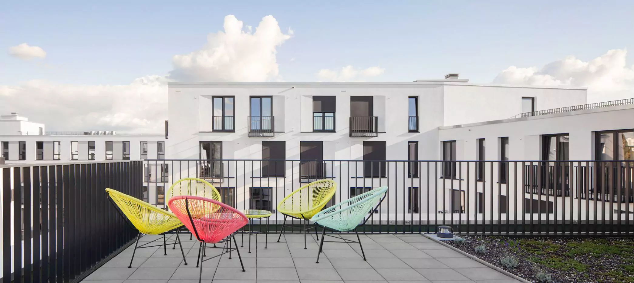 Auf dem Bild ist eine moderne Dachterrasse zu sehen, die mit einem schlichten Stabgeländer von SIMPLUM eingefasst ist. Vor dem Geländer stehen bunte, filigrane Stühle in den Farben Gelb, Rot und Hellblau, die in einem Halbkreis angeordnet sind. Im Hintergrund sind moderne, weiß verputzte Gebäude mit großen Fenstern und französischen Balkonen zu erkennen. Der Himmel ist klar und blau, nur vereinzelt mit ein paar Wolken. Die Szenerie wirkt ruhig und einladend.