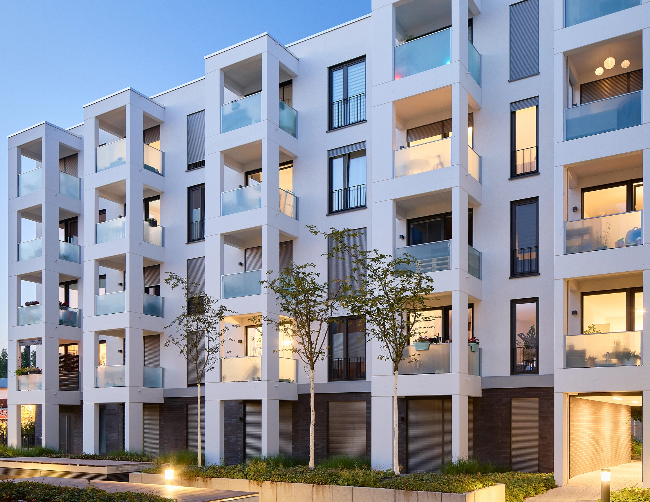 Anfrage erfolgreich  Ein modernes, mehrstöckiges Wohngebäude mit weißer Fassade und vielen Balkonen ist zu sehen. Die Balkone haben Glasbrüstungen und einige sind beleuchtet, was auf bewohnte Einheiten hindeutet. Die Fenster sind überwiegend dunkel oder zeigen ebenfalls Innenbeleuchtung. Im unteren Bereich des Gebäudes ist eine Mischung aus weißem Putz und dunkleren, möglicherweise gemauerten Elementen erkennbar. Vor dem Gebäude befinden sich kleine Bäume und eine gepflasterte Fläche, die von unten beleuchtet wird, was eine warme Atmosphäre schafft. Die Beleuchtung im Inneren des Gebäudes sowie die äußere Beleuchtung lassen vermuten, dass es sich um Abendstunden handelt.